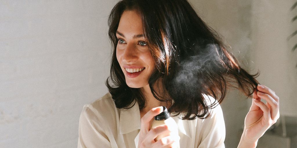 Woman spraying hair product, smiling, indoors, natural light.