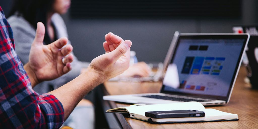 Person gesturing during a virtual meeting, with a laptop displaying a presentation and a smartphone on a notebook, illustrating communication challenges in remote work.