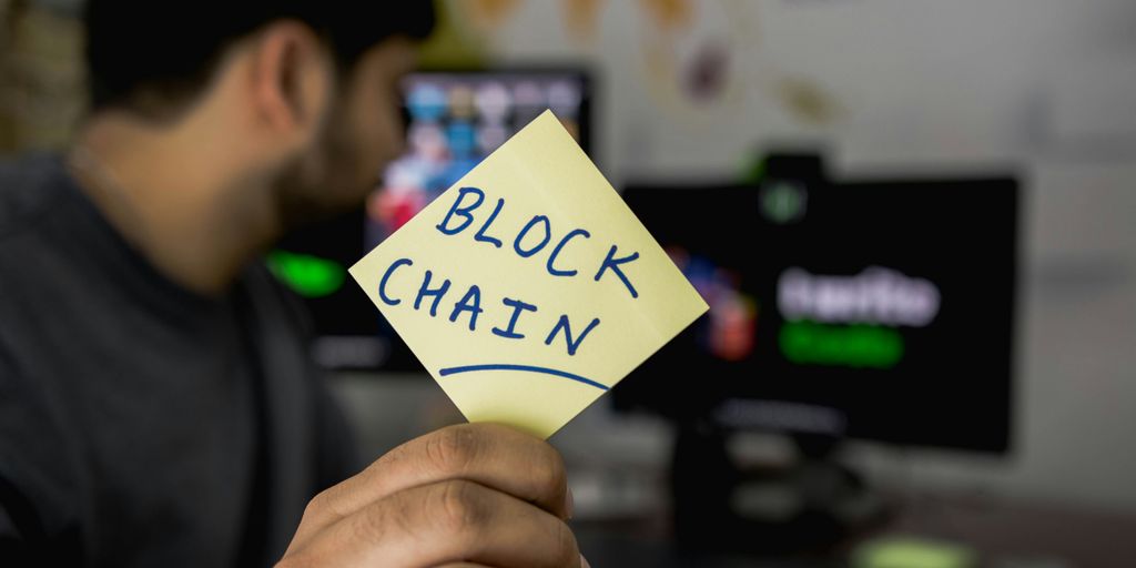 Person holding a sticky note labeled "BLOCKCHAIN" in an office.