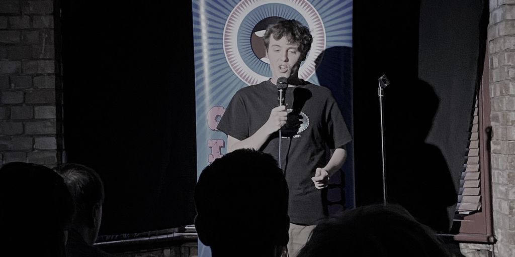 A person performs stand-up comedy on a stage with a banner.