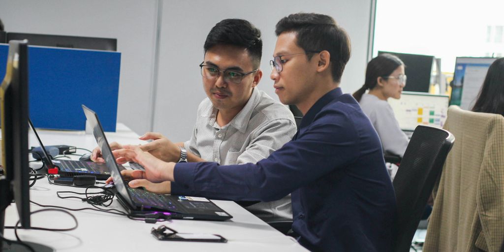 two men sitting at a desk looking at a laptop