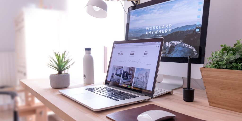 Modern home workspace with laptop displaying productivity metrics, desktop computer with "WORKHARD ANYWHERE" wallpaper, potted plants, and a water bottle, emphasizing effective remote work environment.