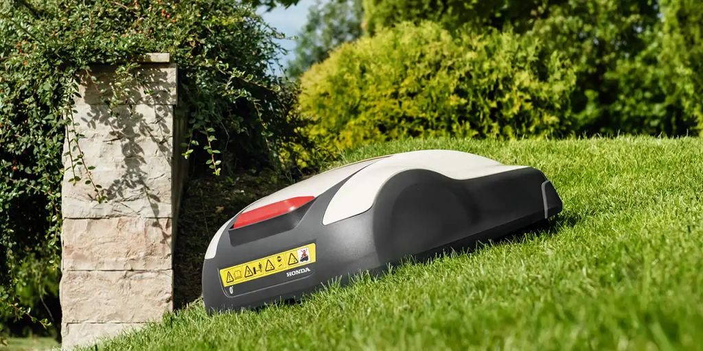 Robot mower on green lawn, surrounded by bushes and trees.