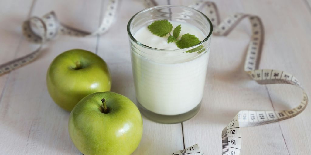 a glass of milk next to two green apples and a measuring tape
