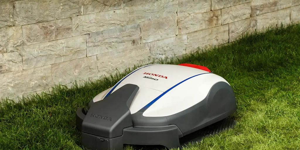 Honda robotic mower resting on green grass by a stone wall.