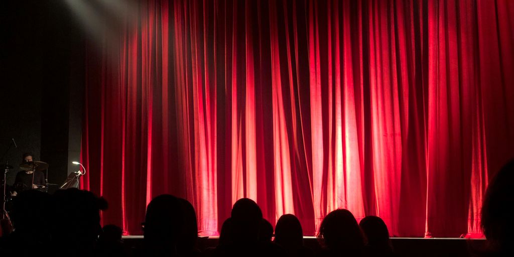 Theater stage with red curtains, audience in shadows waiting.