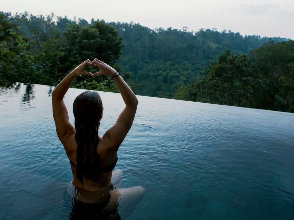 woman in infinity pool making heart hand gesture facing green leafed trees