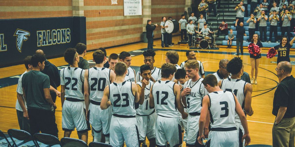 Basketball team huddled together on court, discussing strategy, with fans and cheerleaders in the background, emphasizing teamwork and collaboration in sports.
