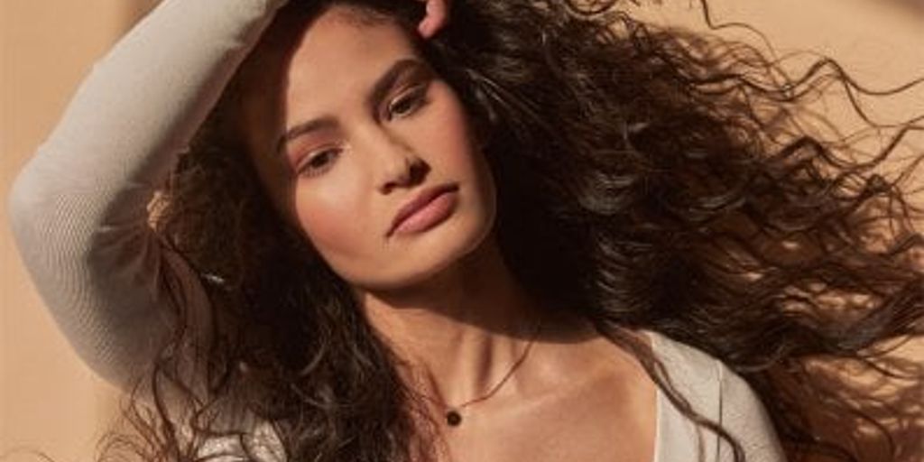 Person posing, long curly hair, subtle light, neutral colors.