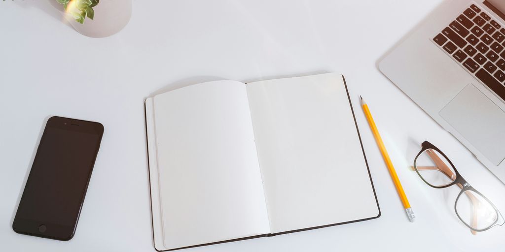 An open empty notebook on a white desk next to an iPhone and a MacBook