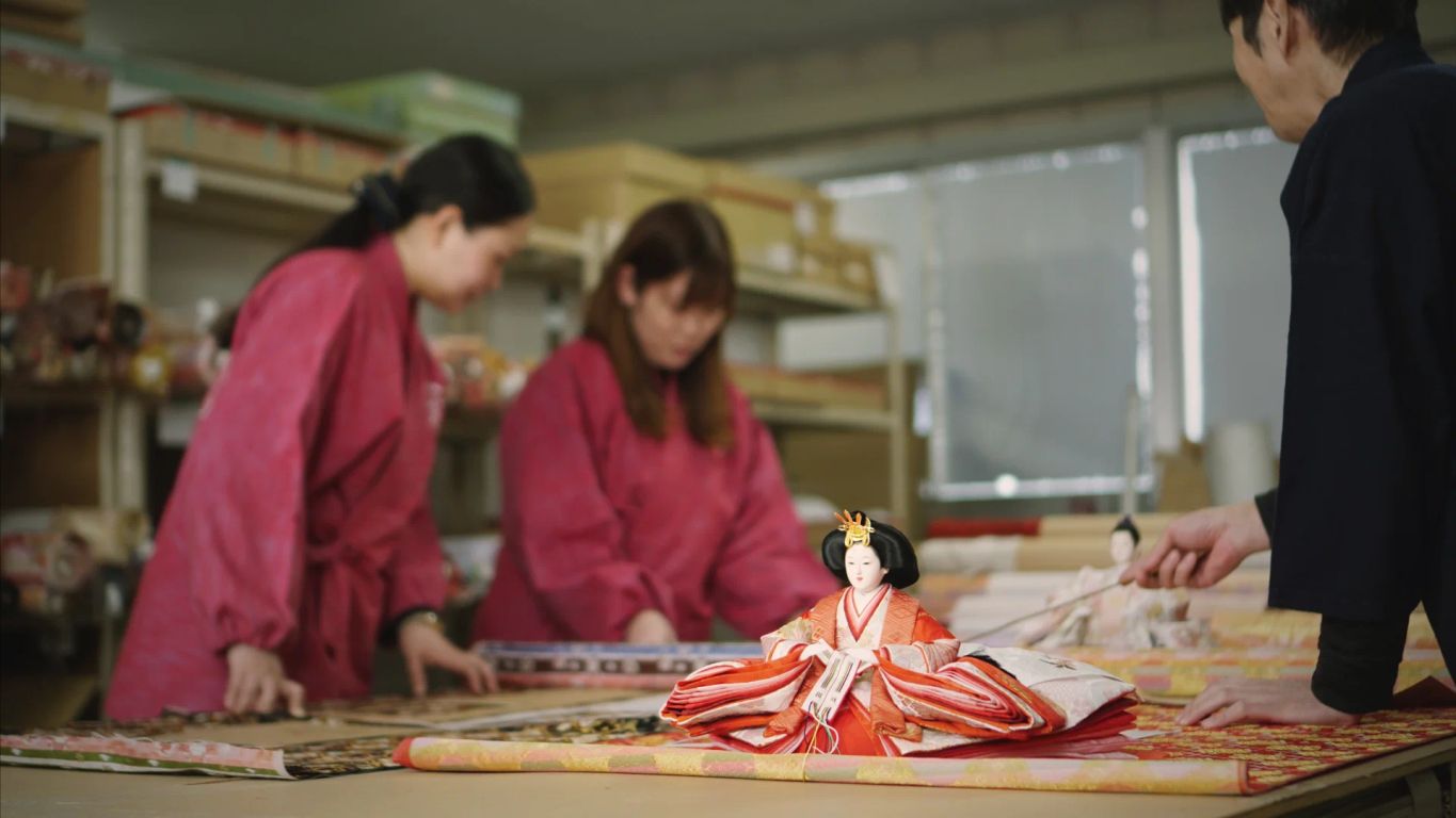 Three individuals in pink clothing work with traditional dolls and fabric.