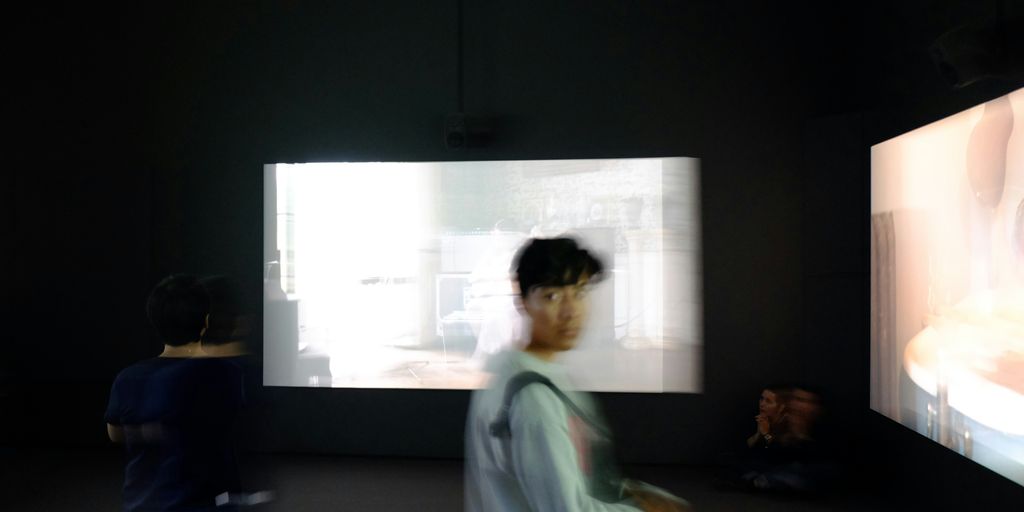 person in grey sweater standing in front large monitors