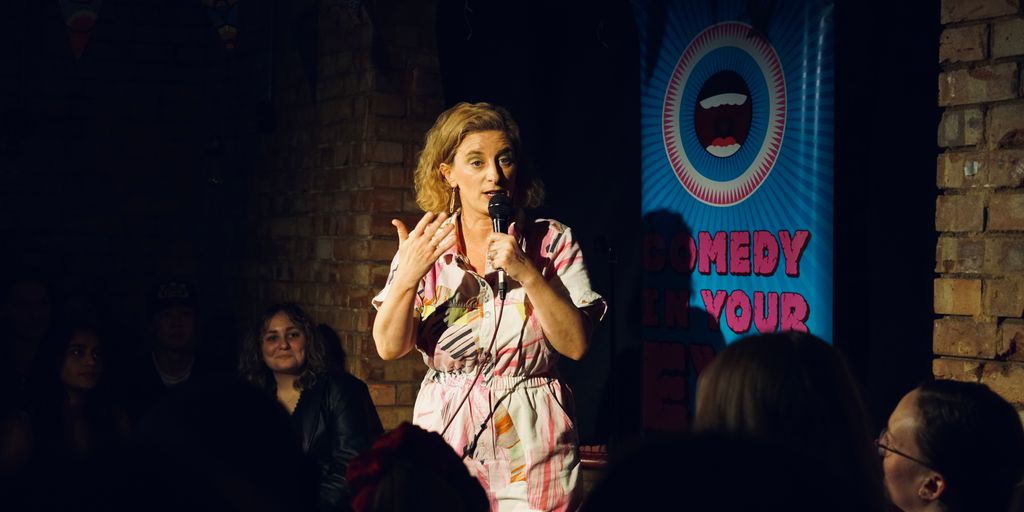 A person performs stand-up comedy in front of a banner.