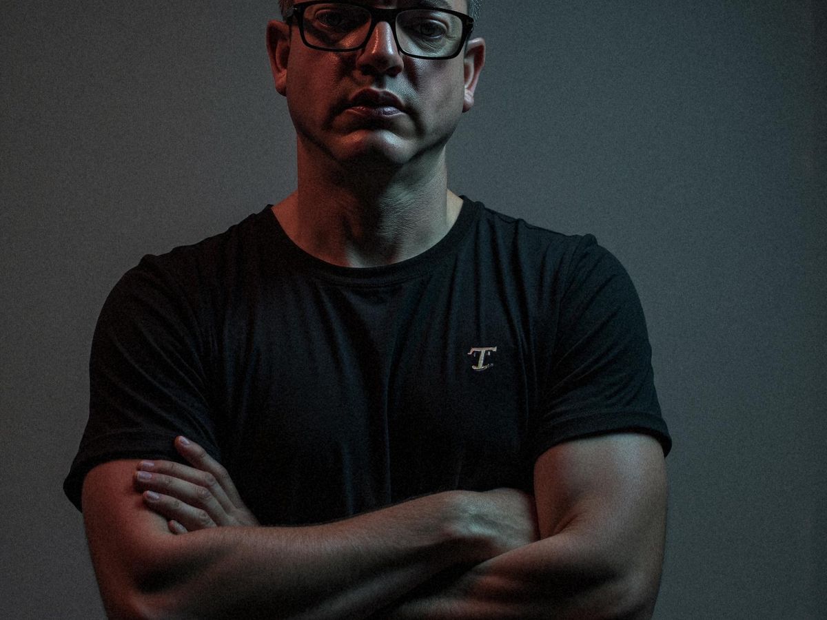 A person stands confidently with arms crossed, in low light.