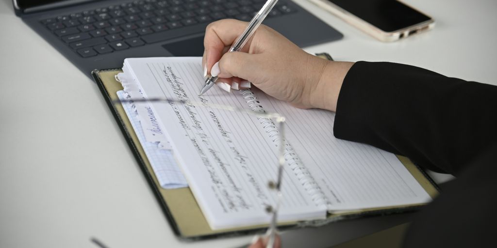 A person writing on a notebook with a laptop in the background